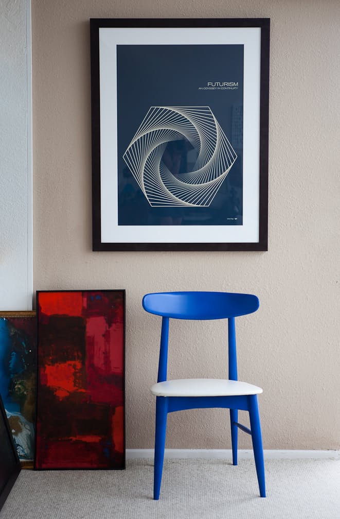 Matte beige wall with a framed geometric poster above a cobalt blue chair and a red abstract canvas