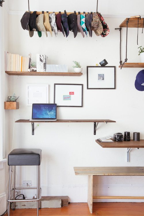 White wall with wood shelves, a wall-mounted laptop desk, framed prints, a bar stool and caps hung from a rod