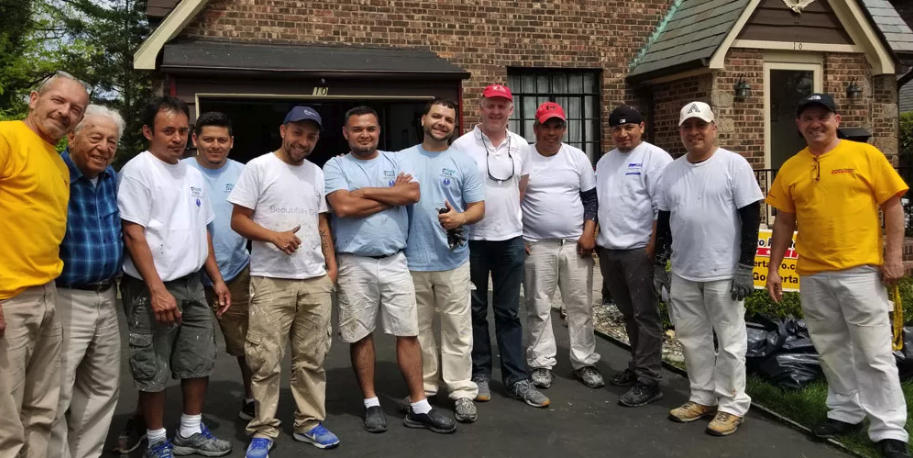 Group of men in work clothes, several in white painter's pants, posing on a driveway in front of a brick house