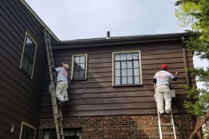Two painters on ladders painting dark brown wood siding on the upper story of a brick house