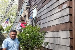 Painter on a ladder working on brown wood siding spotted with gray primer, with a crew member in front