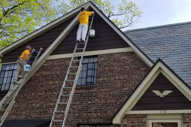 Two painters in yellow shirts on tall ladders painting the dark brown gable of a brick house