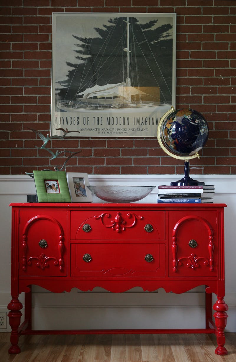 Antique sideboard painted glossy red against a brick wall, with a sailboat poster and a desk globe