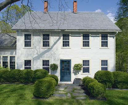 White clapboard farmhouse with a teal front door, rounded boxwood shrubs and a stone path
