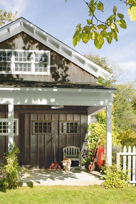 Weathered board and batten cottage with white trim, a covered porch, a bench, a red wheelbarrow and a dog