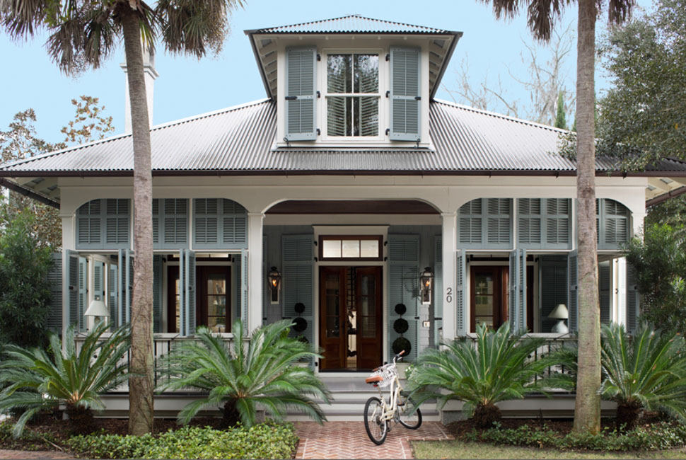 White coastal house with blue-gray shutters, a metal roof with a dormer, a front porch and palm trees