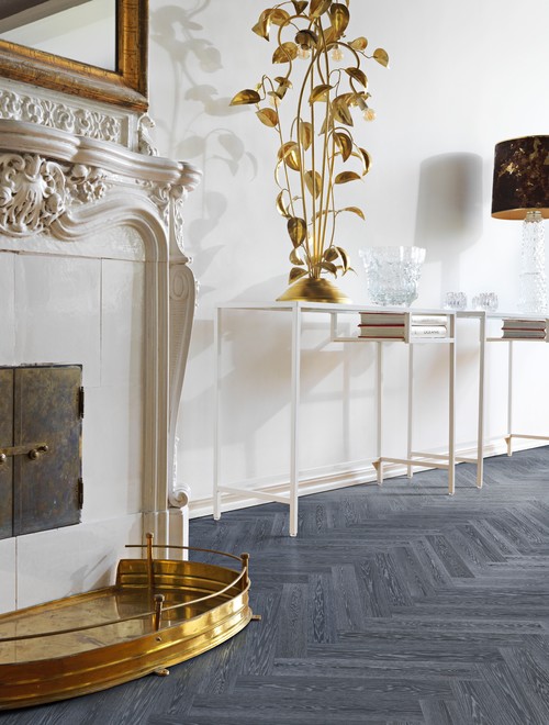 Dark gray herringbone wood floor by a carved white fireplace with a brass fender and white console tables
