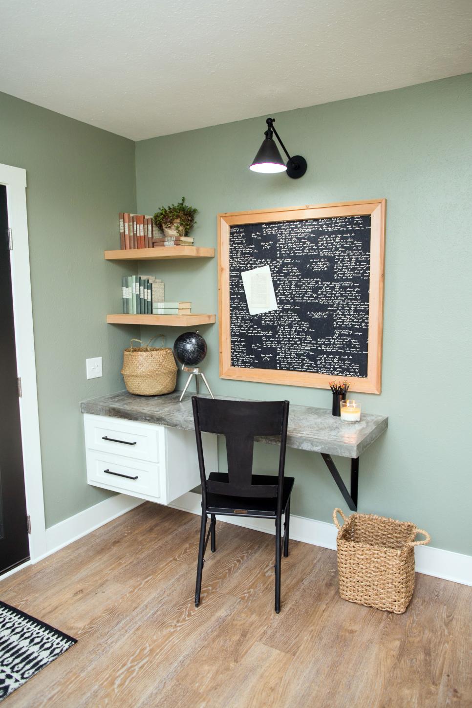 Home office nook with sage green walls, a floating concrete-look desk, wood shelves and a black framed pinboard