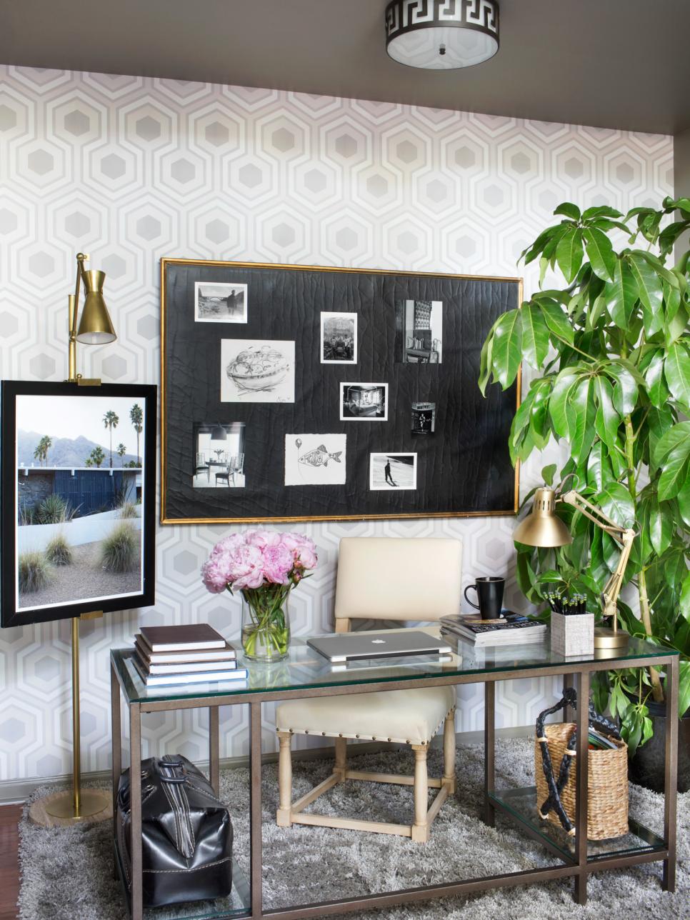 Home office with gray hexagon wallpaper, a glass desk, a gold-framed black pinboard, brass lamps and a large plant