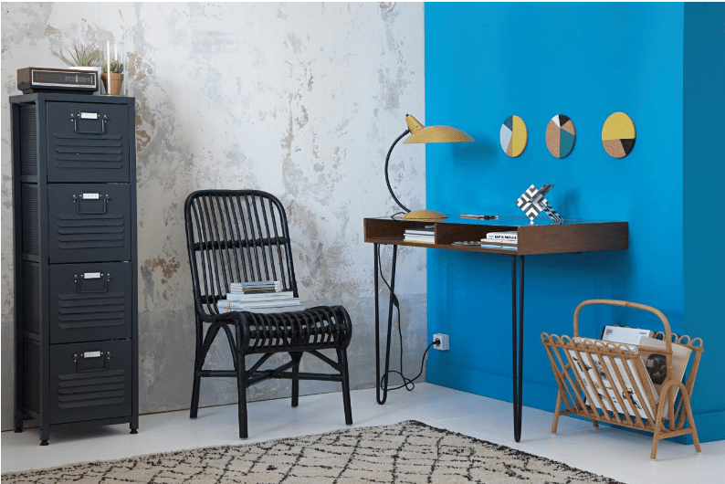 Home office corner with a bright blue accent wall, a slim wood desk, yellow lamp, black rattan chair and metal cabinet