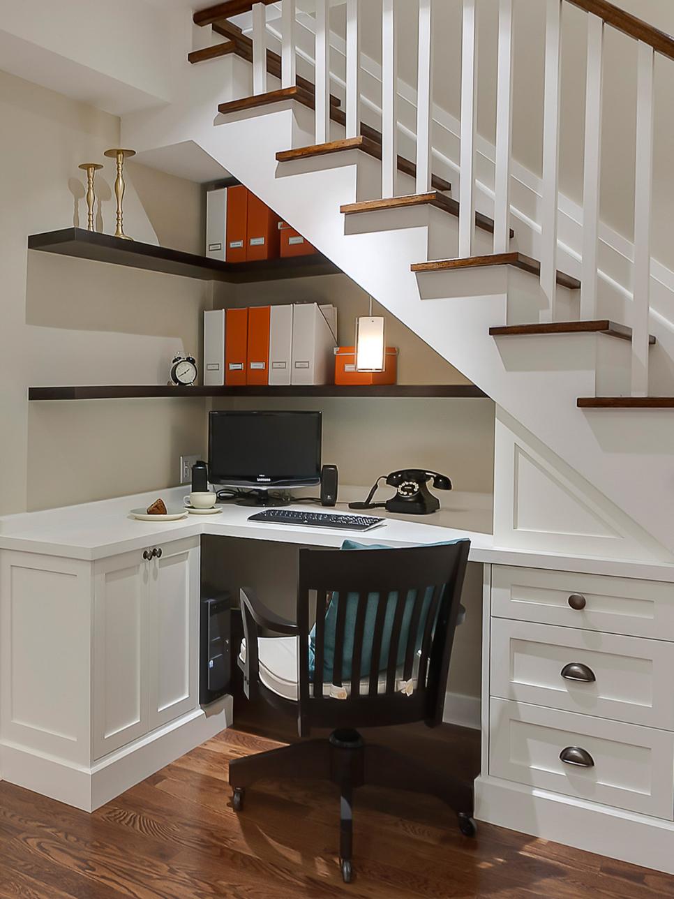 Home office built under a staircase with a white corner desk, drawers, dark floating shelves and a black chair