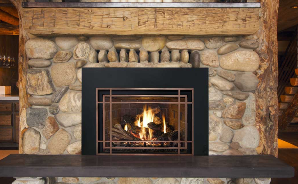 River rock fireplace with a rustic wood beam mantel and a black gas insert with a lit fire