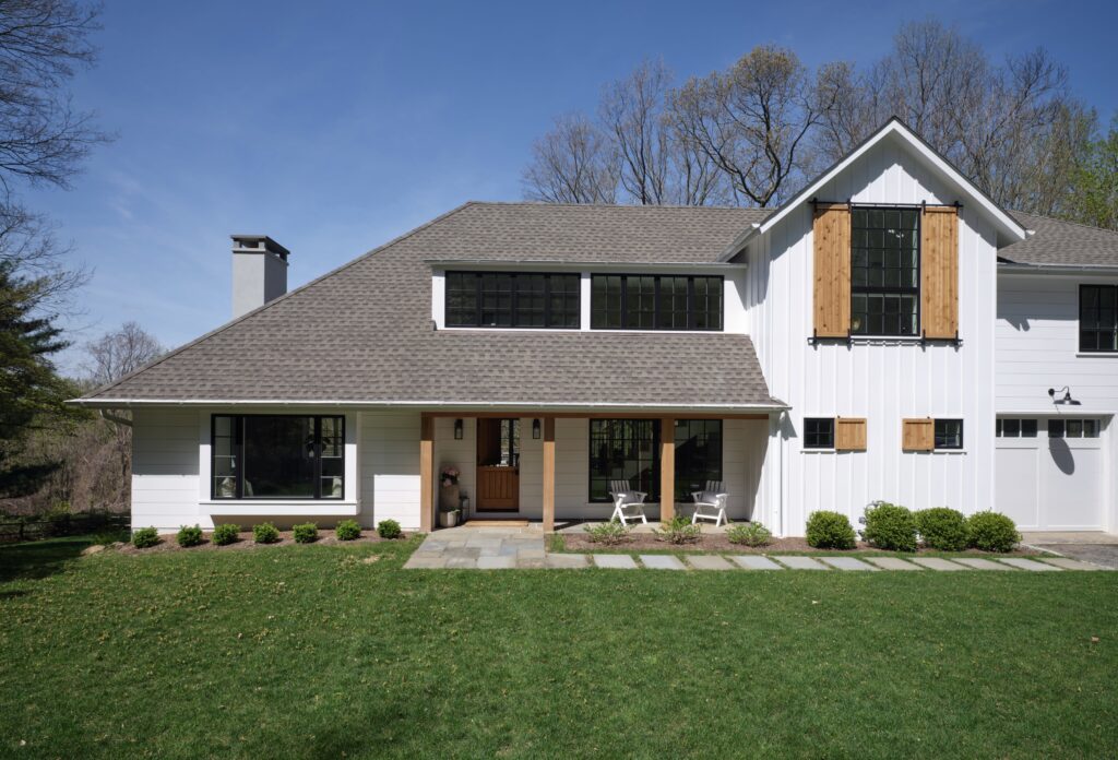 White board and batten farmhouse with a gray roof, natural wood shutters, a covered front porch and a green lawn