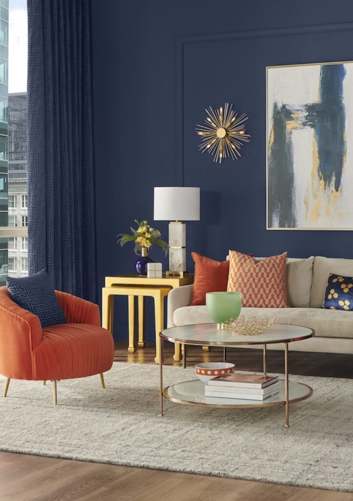 Living room with navy blue walls, an orange armchair, a cream sofa, a yellow side table and gold starburst wall decor