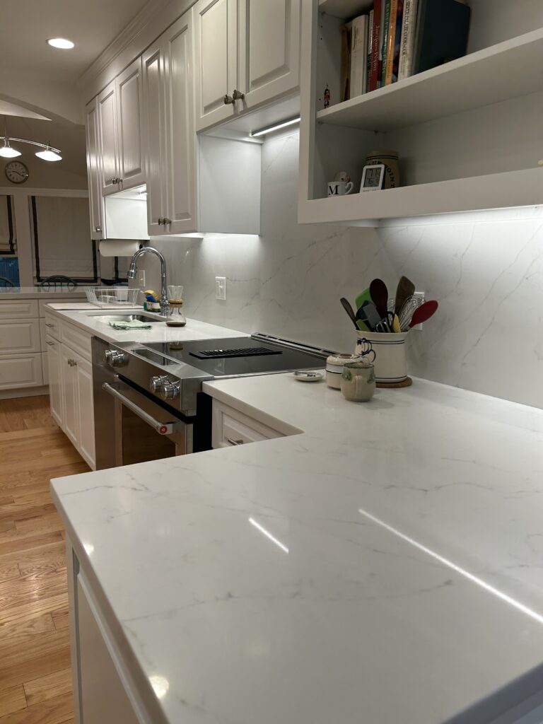 White kitchen with white cabinets, marble-look counters, under-cabinet lights and an open shelf with books