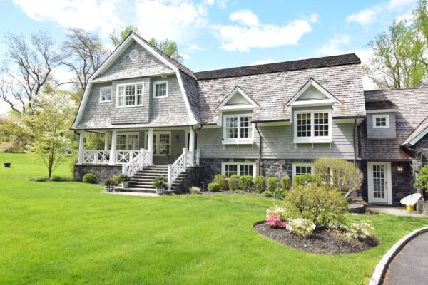Large gray shingled house with a stone foundation, white porch railings, a green lawn and planted beds