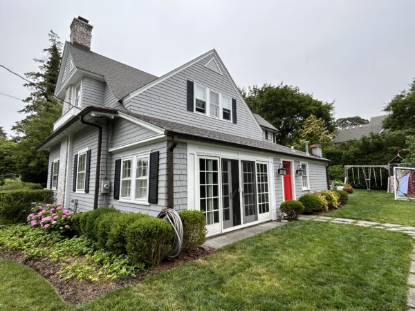 Gray shingled house with black shutters, a red door, white French doors, shrubs and a green lawn