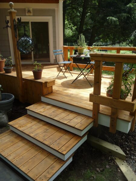 Natural wood deck with wide steps, railings, potted plants and a small table with folding chairs