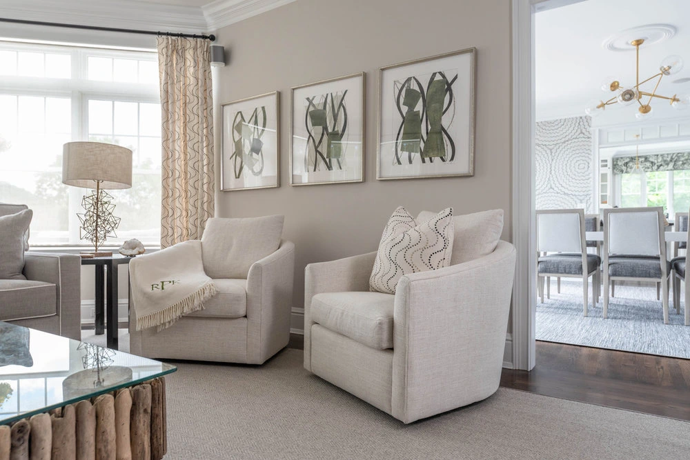 Sitting area with two cream armchairs, three framed black and white prints on a greige wall and a glass coffee table